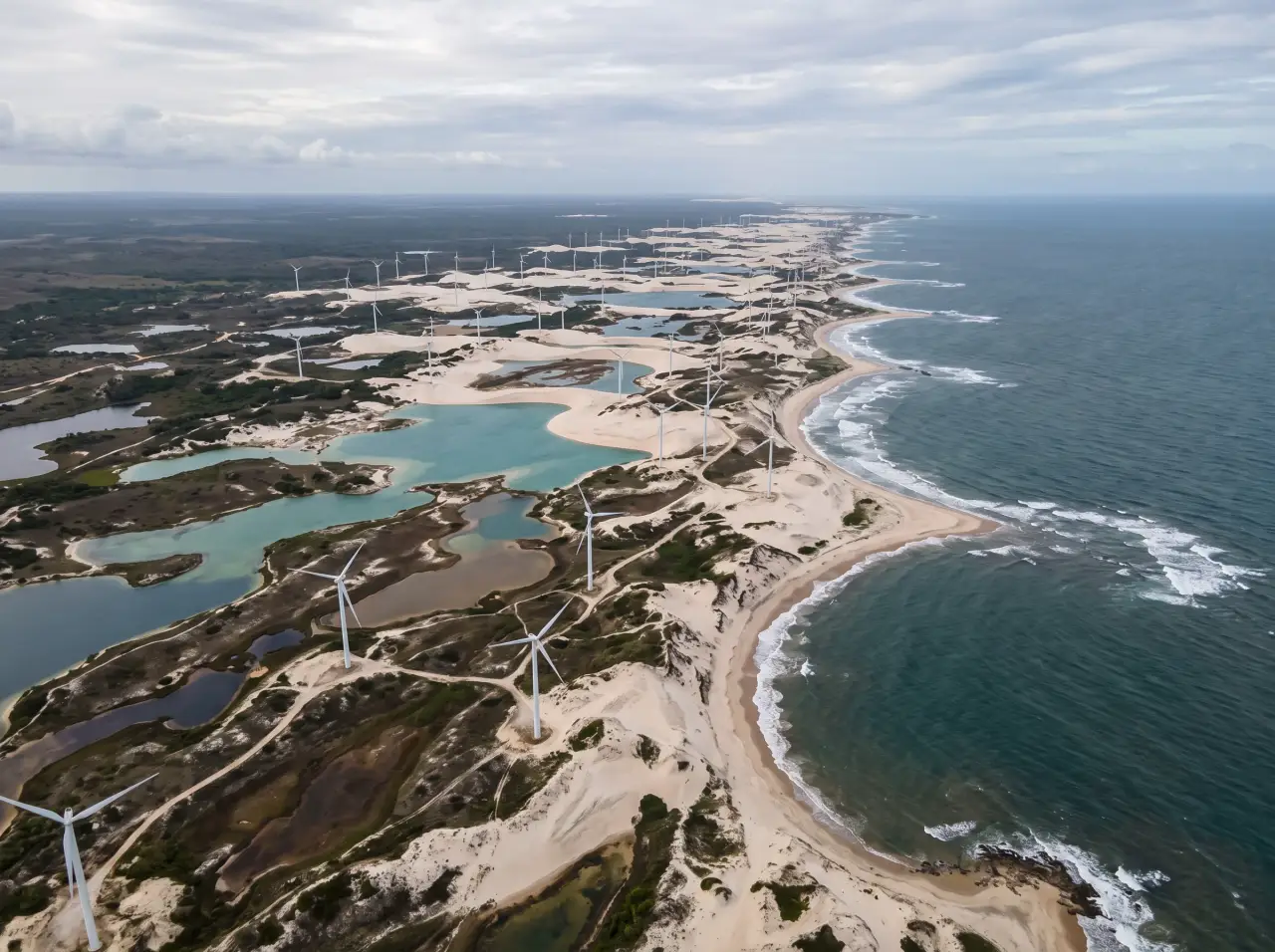 Vista aérea de um parque eólico no litoral do Nordeste, com turbinas sobre dunas e lagoas.